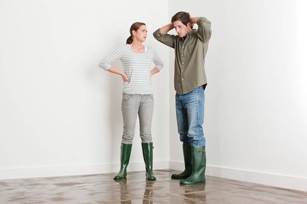 Two people standing in a bare room, one gesturing while the other listens, both wearing green boots.