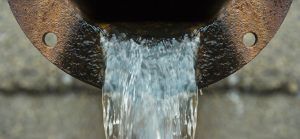 Water pours from a rusted metal pipe with bolt holes.
