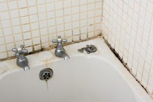 Dirty bathtub corner with old faucets, drain, and stained tiled wall