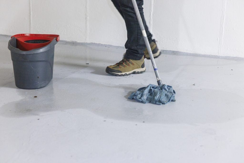 Person mopping a white floor beside a gray bucket with a red cloth cover