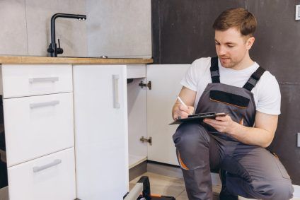 Man in a white shirt kneeling by an open cabinet, writing on a clipboard in a kitchen under a sink
