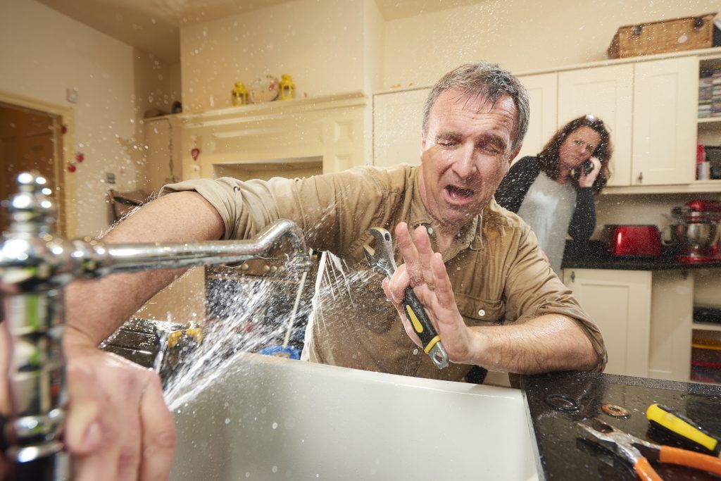 Man splashed by water in a kitchen sink, with a woman smiling in the background