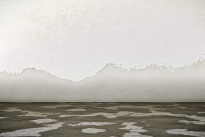 Foggy mountains beyond a gray, rippled shoreline under a pale sky
