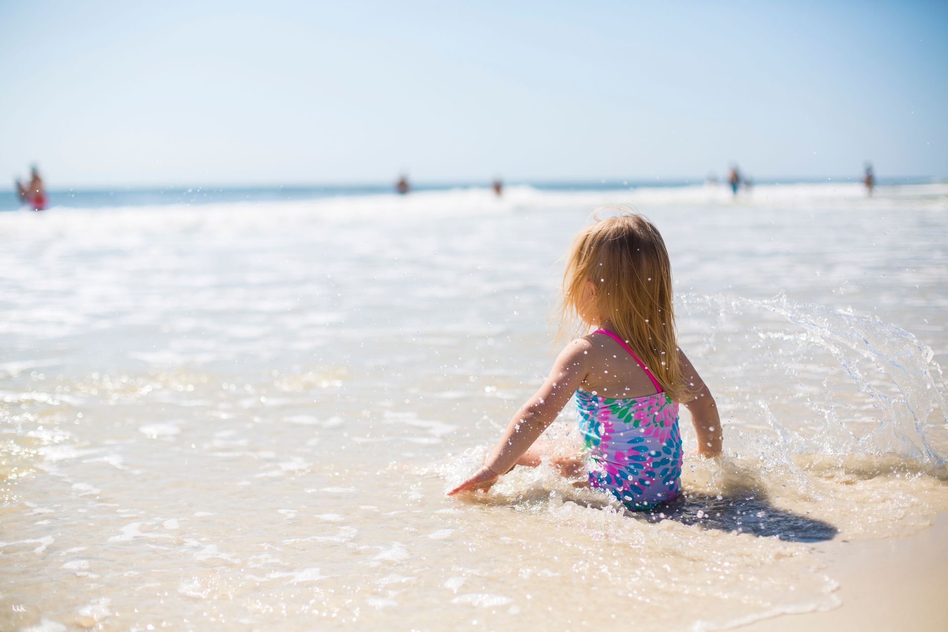Child sitting in shallow ocean water at a sunny beach, wearing a colorful swimsuit.