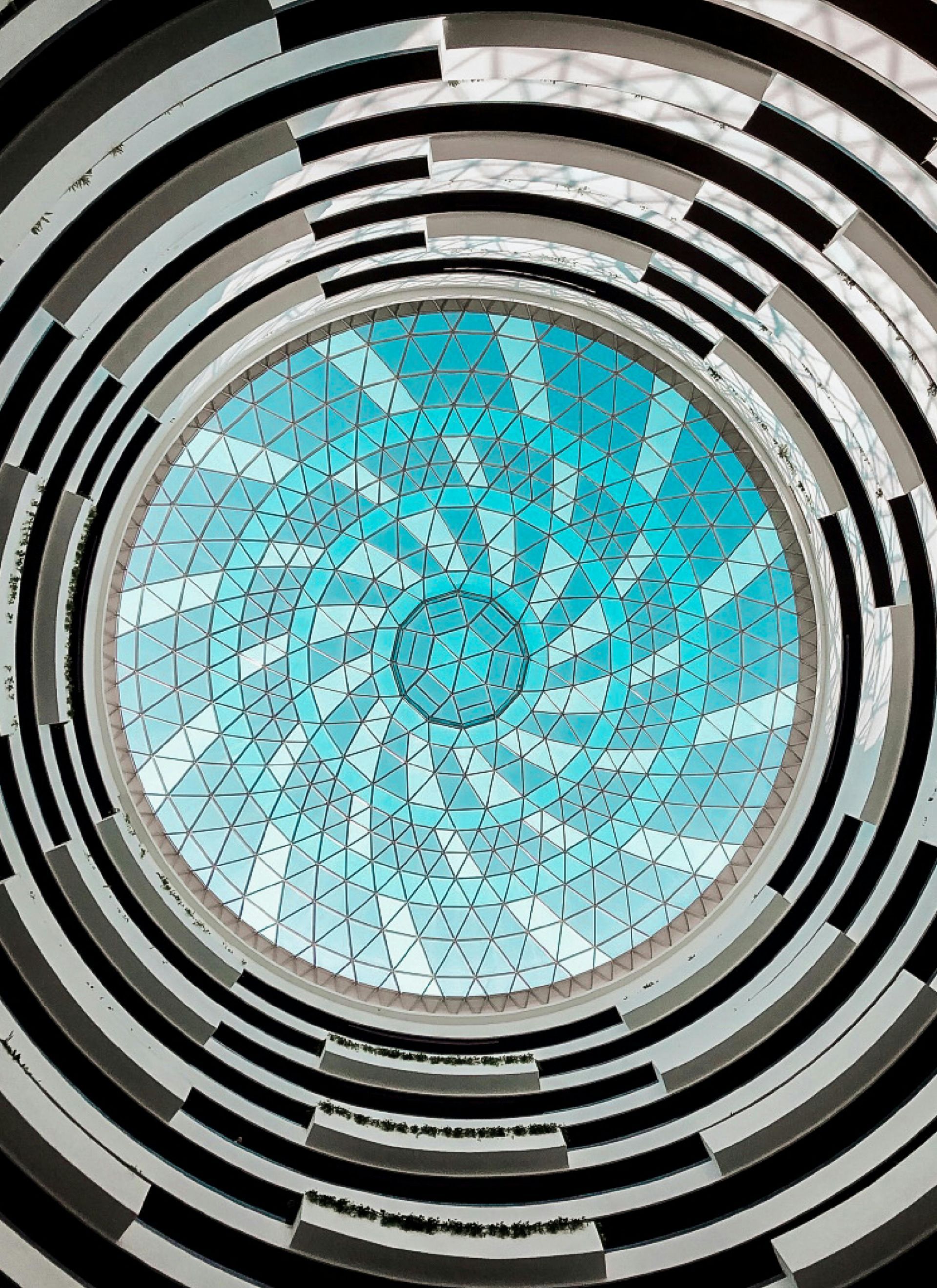 מבט כלפי מעלה דרך אדריכלות קונצנטרית ומעגלית, אל חלון גג בגוון כחול ובדוגמא גיאומטרית.