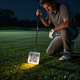 A golfer kneeling on a dark course, inspecting a glowing square QR code marker stuck into the grass.