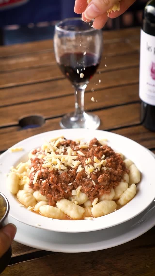Pasta con salsa de carne en un plato blanco, con una mano rallando queso y una copa de vino tinto cerca.
