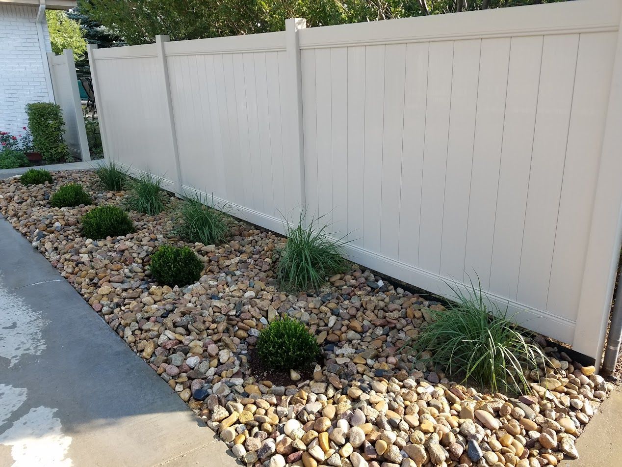 Rock-covered garden bed with small shrubs beside a white fence and sidewalk