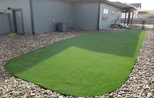 Artificial turf strip beside a gray house, bordered by rocks.