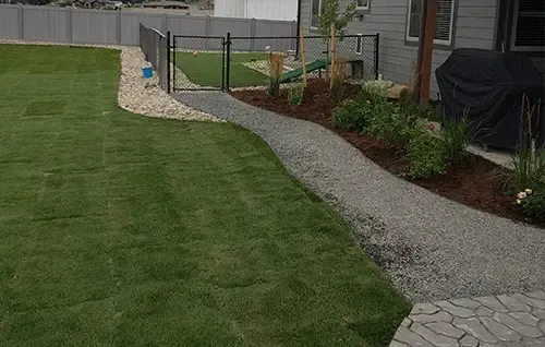 Backyard with a green lawn, gravel path, garden beds, and a fenced gate near a house.