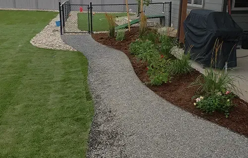 Gravel path beside a green lawn, curving past flower beds and a fenced patio area with covered grill