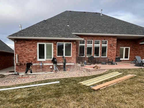Backyard with red brick house, workers on patio, and lumber laid on the grass
