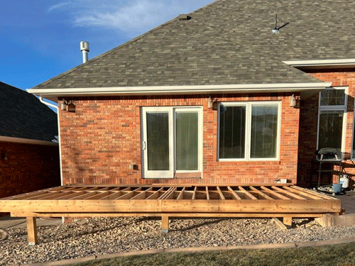 Backyard deck under construction beside a brick house with sliding glass doors and gravel ground