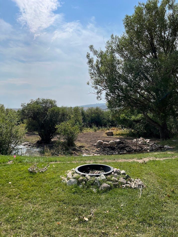 Grassy yard with a fire pit, shrubs, and trees under a partly cloudy sky