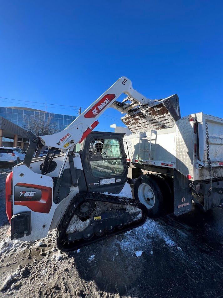 Bobcat skid steer dumping snow into a truck under a bright blue sky