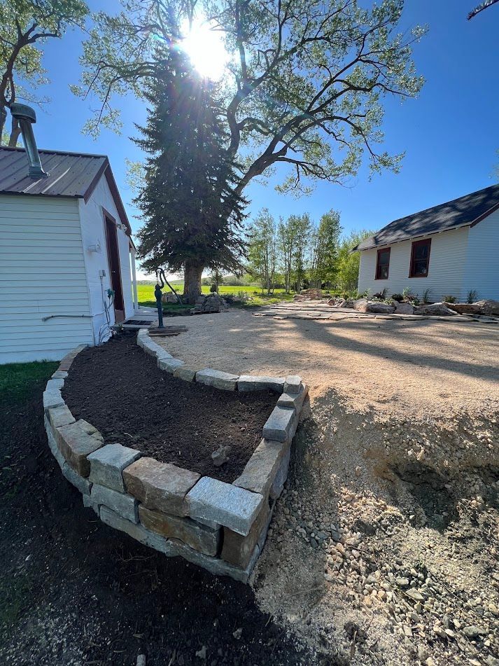 Stone planter bed in a sunny yard between two white houses, with a large tree and gravel ground.