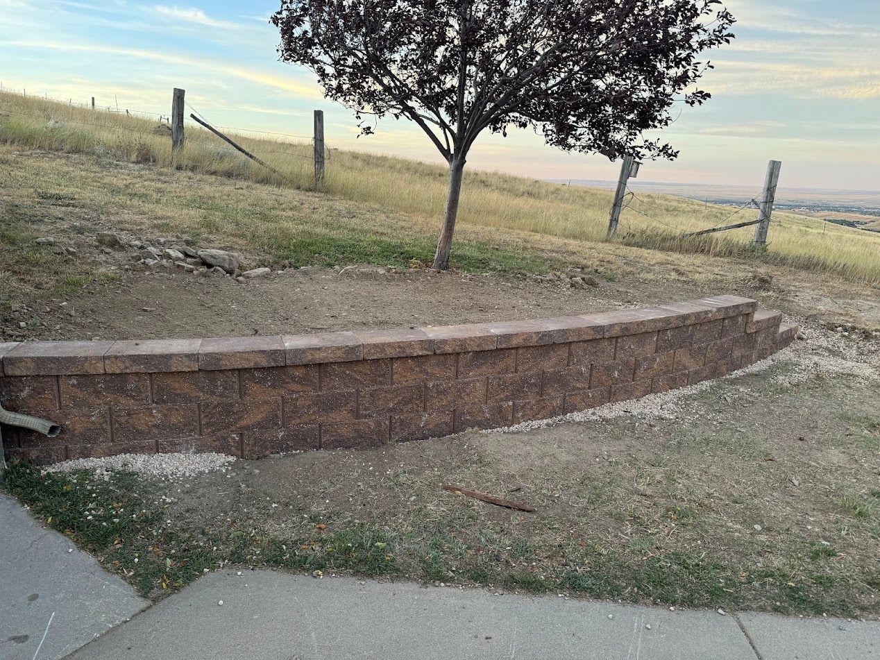Curved brick retaining wall beside a tree on a grassy roadside slope near a fence