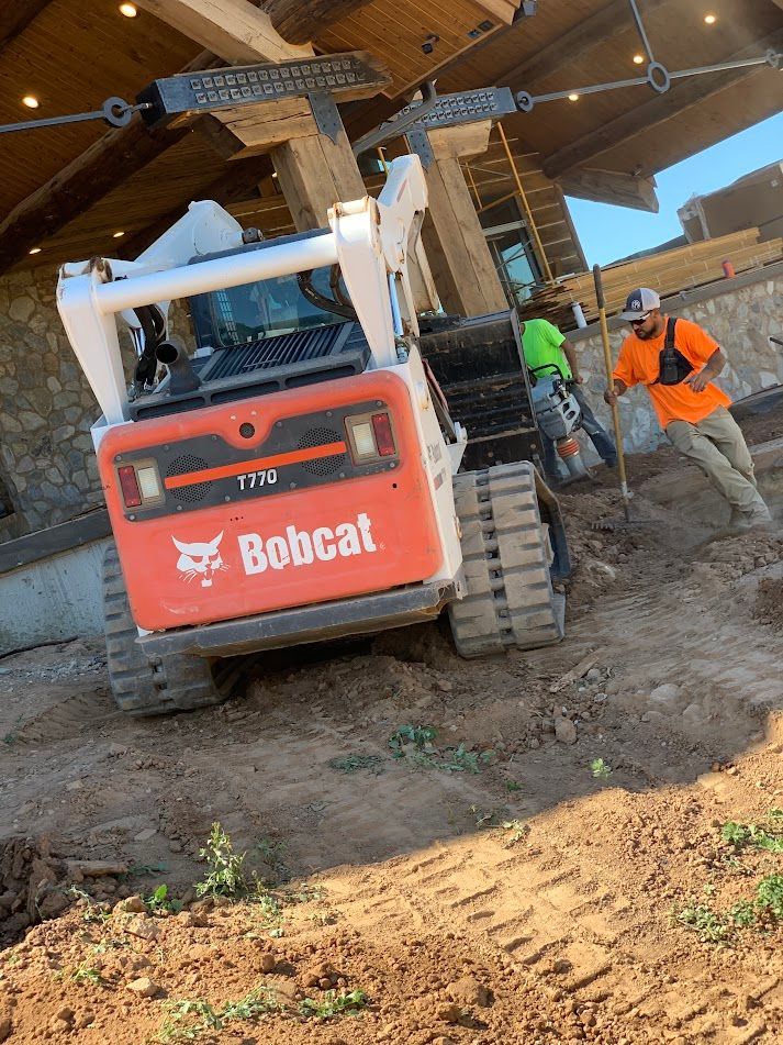 Bobcat skid-steer with two workers on a muddy construction site under a wooden structure