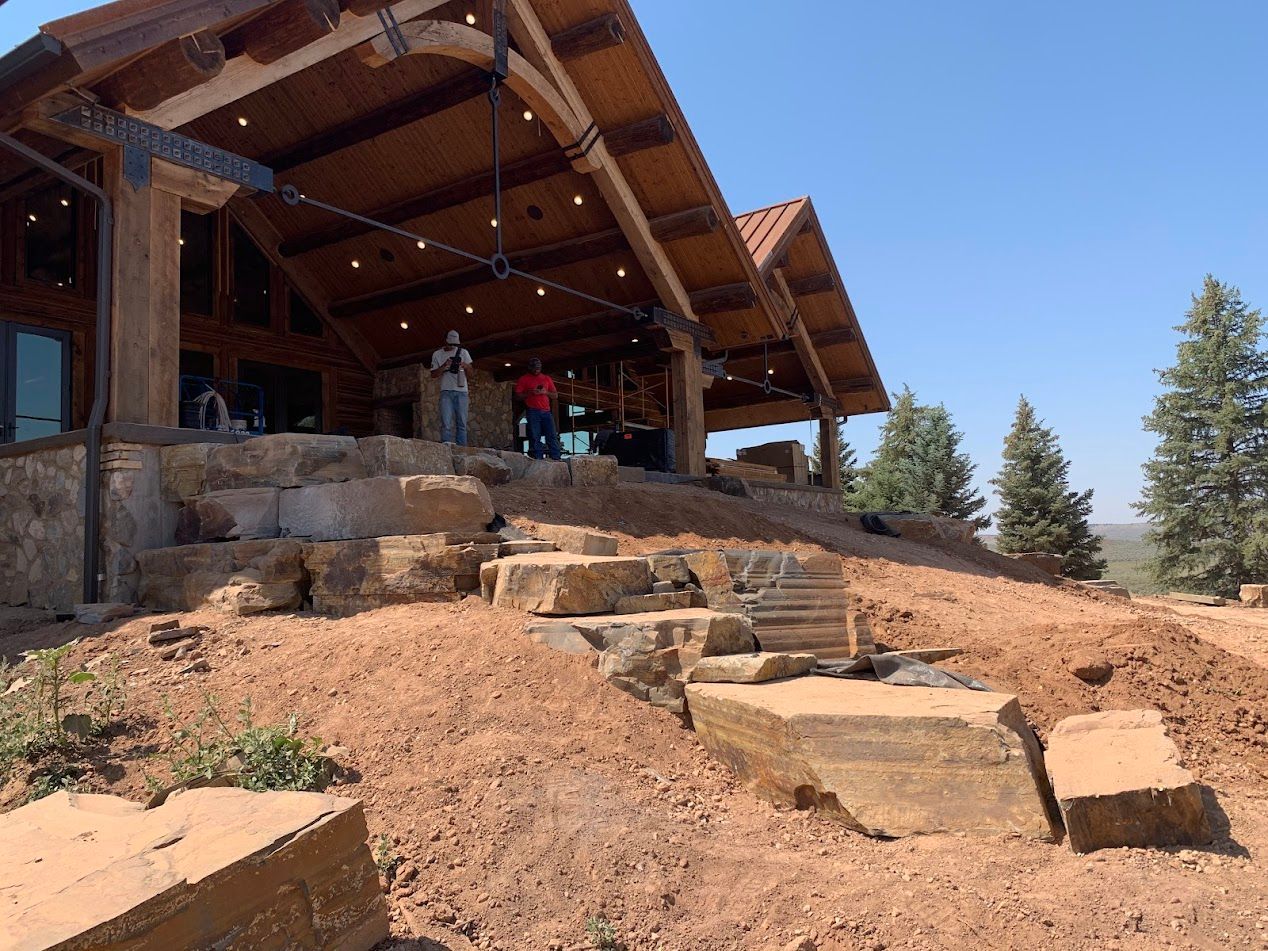 Rustic stone-and-wood house with a covered porch on a hillside under a clear blue sky
