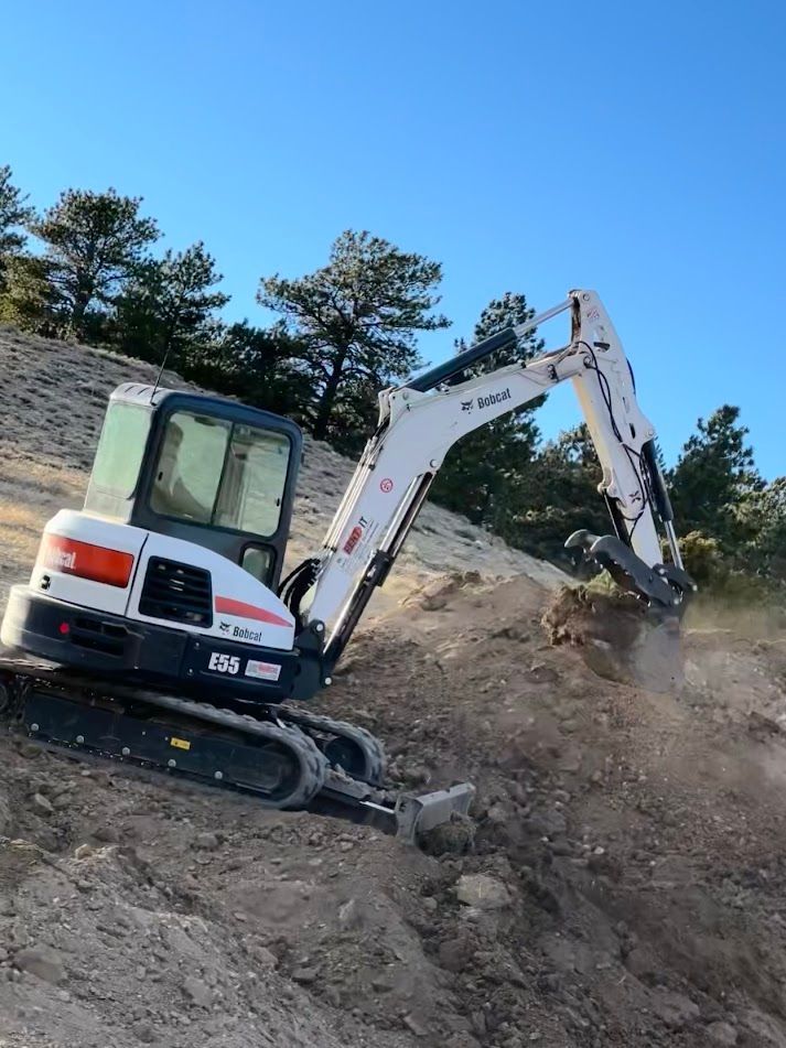 Excavator digging a dirt slope beside a tree line under a clear blue sky
