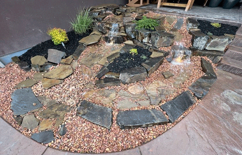 Stone garden fountain with water flowing over black rocks and pink gravel border