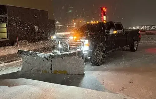 Pickup truck plowing snow at night in a snowy parking lot, with headlights and amber beacon lights on.