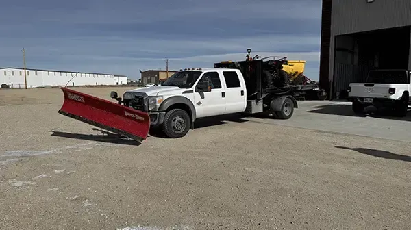 White pickup truck with a red snowplow attachment parked beside a building in a gravel lot