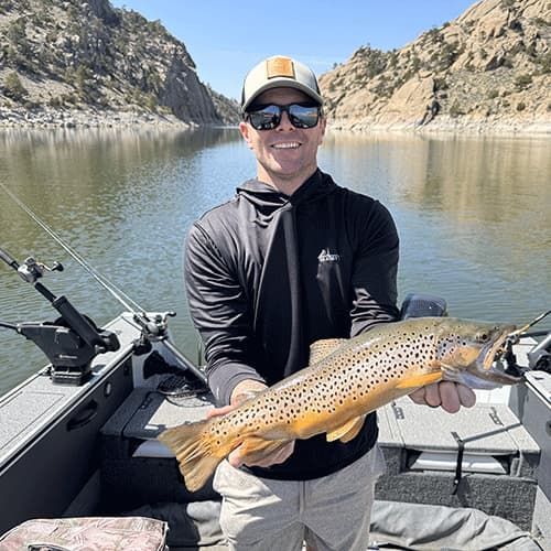 Angler smiling in a boat, holding a spotted fish on a calm lake between rocky cliffs