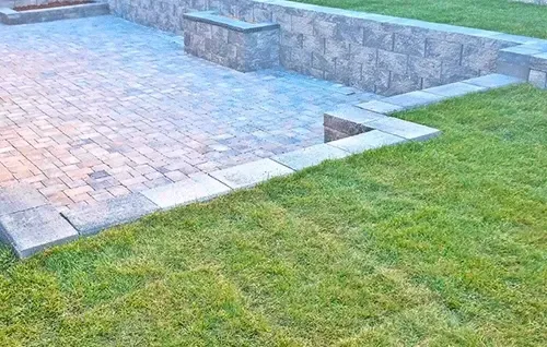 Stone patio beside green lawn with low retaining wall and steps in a landscaped yard