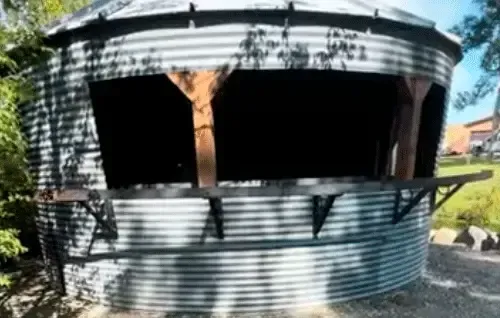 Corrugated metal shed with large open windows, dark interior, and a tree trunk against the front wall