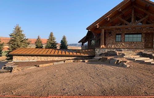 Rustic log cabin with stone steps and a landscaped dirt yard under a clear sky.