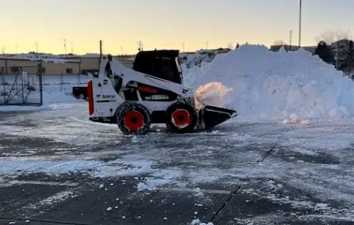 Red-and-white skid steer clearing snow in a parking lot beside a large snow pile at sunset