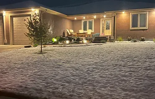 Snowy front yard with a warmly lit house and garage at night