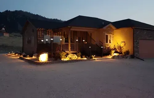 Warmly lit house exterior at dusk with glowing yard lights and mountains in the background