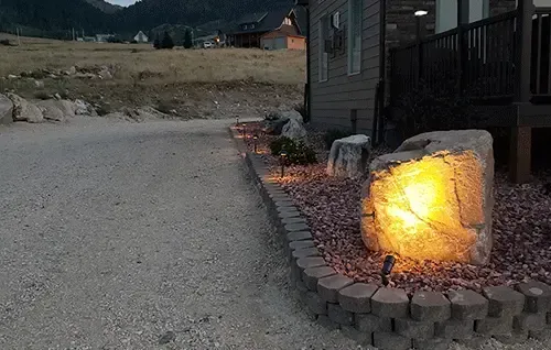 Gravel driveway beside a house, with a glowing rock garden light along a landscaped border at dusk.