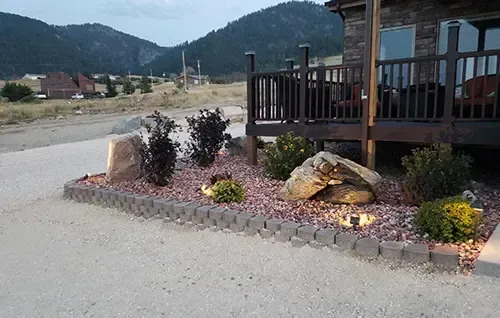 Rock garden with flowers and a wooden porch beside a paved road, with mountains in the background