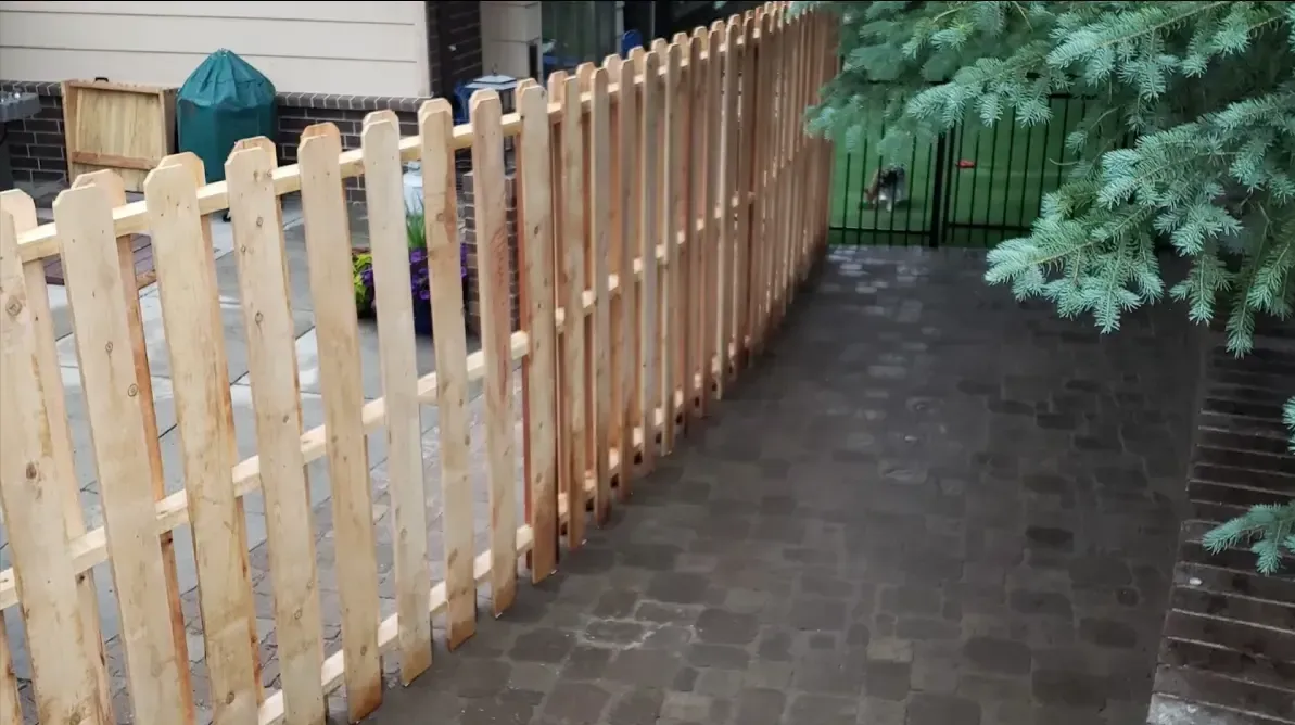 Wooden fence beside a brick walkway in a narrow garden path with evergreen trees.