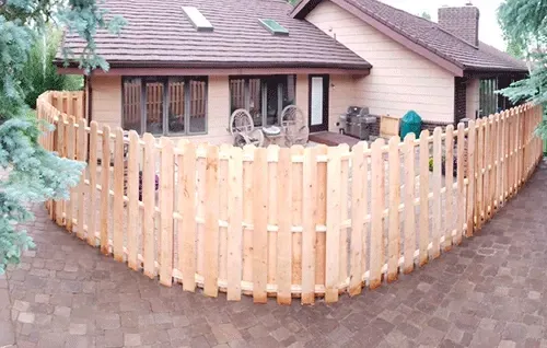 Curved light wood picket fence around a brick house with a brown roof and paved driveway
