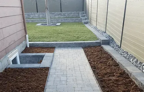 Narrow paver walkway between buildings leading to a grassy backyard, with raised garden beds on both sides