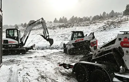 Excavators and a skid steer clearing snow at a muddy, snowy construction site.