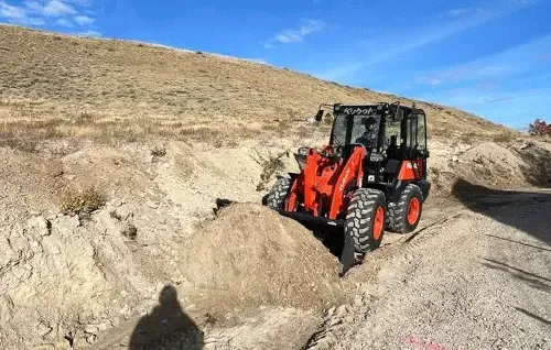 Red skid steer digging into a dirt embankment beside a gravel road in a dry hillside landscape.