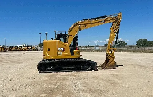 Yellow CAT excavator on a dirt lot with its bucket raised, under a clear blue sky.