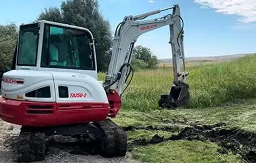White and red mini excavator digging a muddy ditch beside a grassy field under a clear sky
