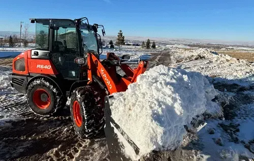 Orange tractor clearing a large snow pile on a snowy roadside under a clear blue sky