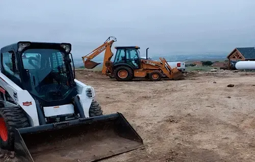 Two construction vehicles on a dirt lot with a beach and ocean in the background