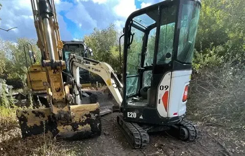 Two excavators working in a muddy wooded area, one white mini digger in the foreground.