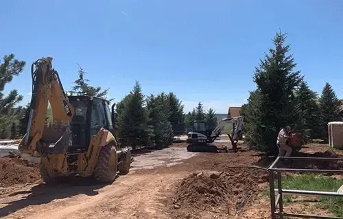 Backhoe and dump truck on a dirt road at a construction site with trees and clear blue sky