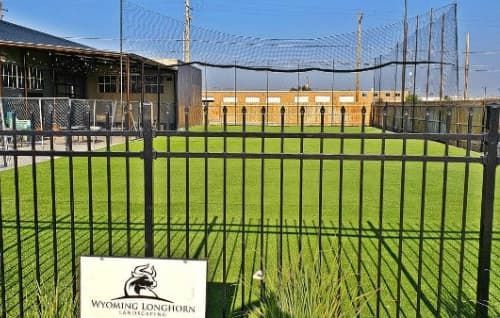Baseball field behind black fence with green turf and buildings in the background