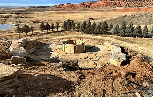 Ancient circular stone ruin in a dry desert landscape with trees and red cliffs in the background