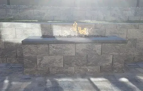 Stone bench with a small flame on top against a gray stone wall, sunlight casting shadows on the ground